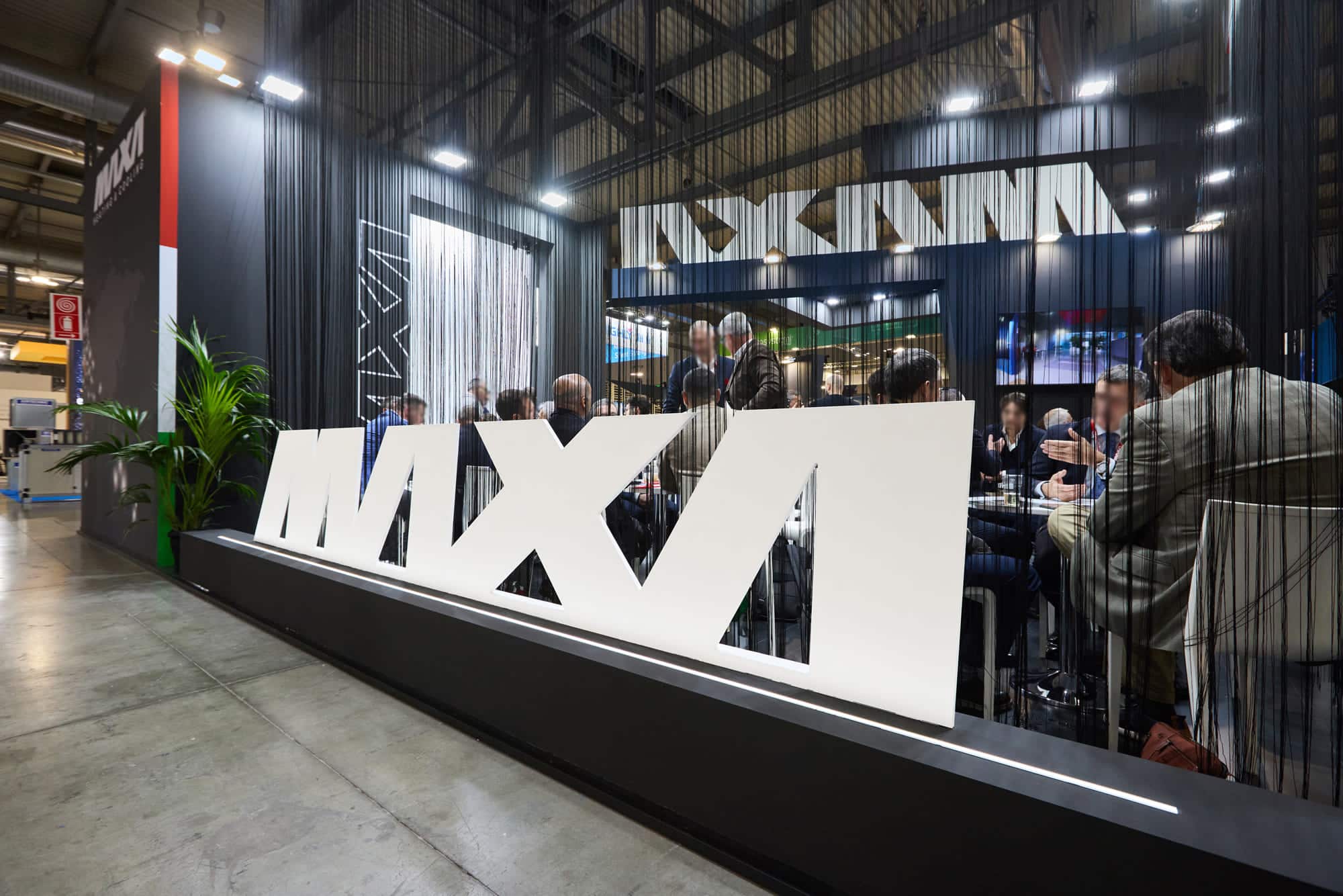 Dettaglio del logo Maxa in 3D bianco con tenda a fili nera e area meeting sullo sfondo allo stand MCE Milano.