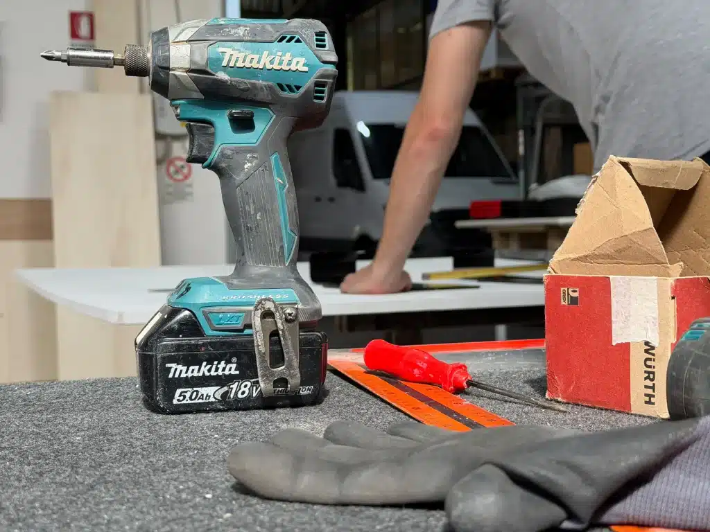 Avvitatore a batteria, guanti da lavoro e attrezzi in primo piano su un banco. Sullo sfondo sfocato, un artigiano prende le misure su un pannello all'interno della falegnameria.